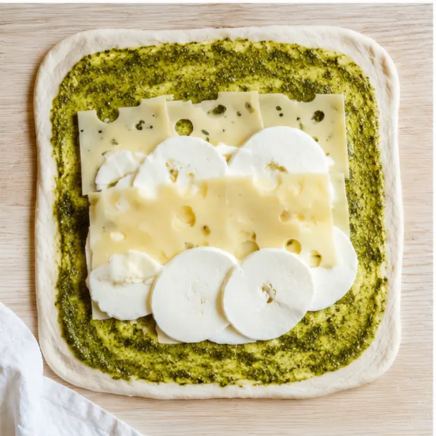 Spreading basil pesto and layering cheese on the rolled rectangle
