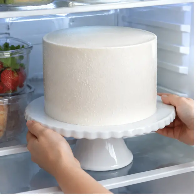 Step 1: Chilling the fully frosted plain white buttercream layer cake on a white cake stand inside the refrigerator until the frosting feels firm